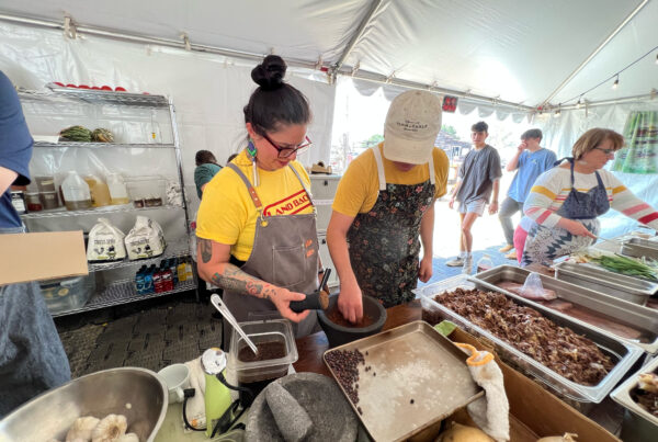 Two chefs work at a table covered in dishes filled with a variety of ingredients. They're in what appears to be a tent structure. One of the chefs is grinding chile peppers in a molcajete.