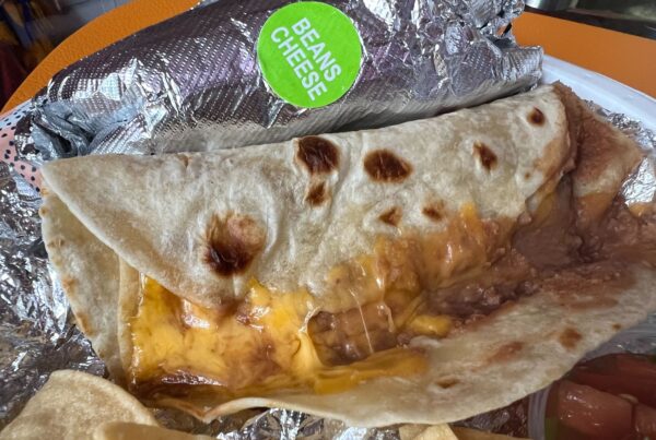 A flour tortilla filled with refried beans and melted cheese sits on top of the foil in came in on a plate. Some chips and a small cup of pico de gallo are seen off to the side.