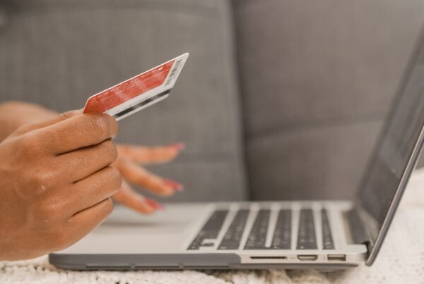 A photo of the hands of a person hovering over a laptop. One hand holds a credit card, suggesting the person is shopping online.