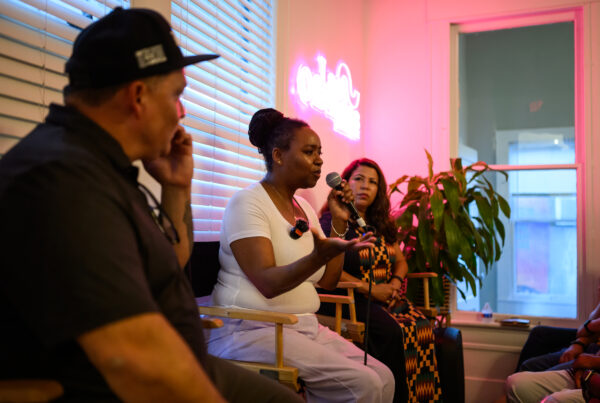 Three people sit in chairs facing the crowd in front of them. In the middle a woman holds up a microphone and is speaking while the other two look on. The woman is Windy Goodloe.