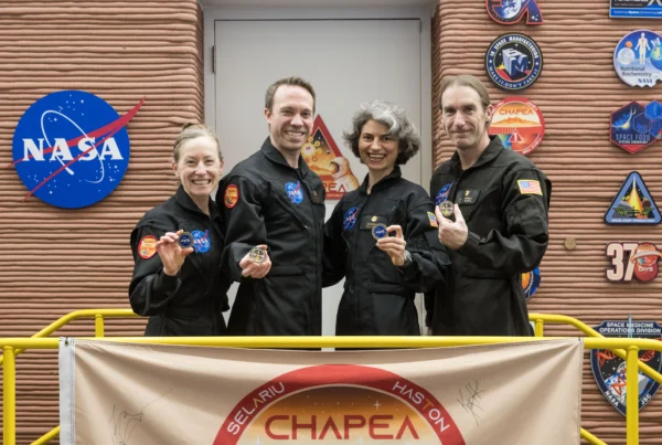 Four people wearing black NASA work suits smile and hold up medallions outside of a 3D printed enclosure in which they spent more than a year.