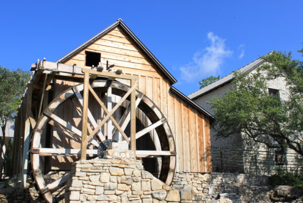 A modern photo shows the replica wooden and stone mill with its big wheel in front of a bright blue sky.