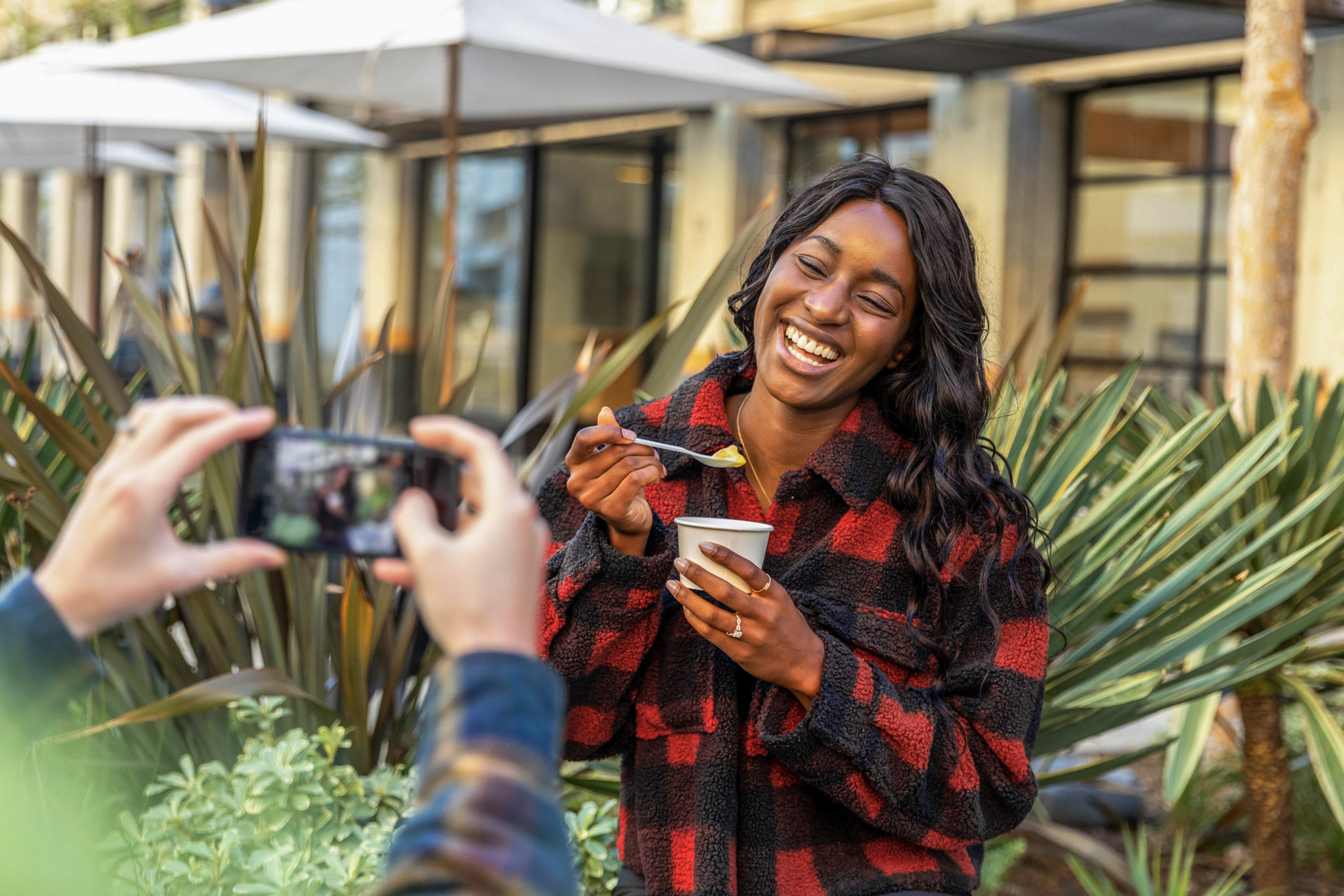 a stock photo of a young women being photographed by another person with a smartphone, while standing outside a building while posing with a small bowl of food