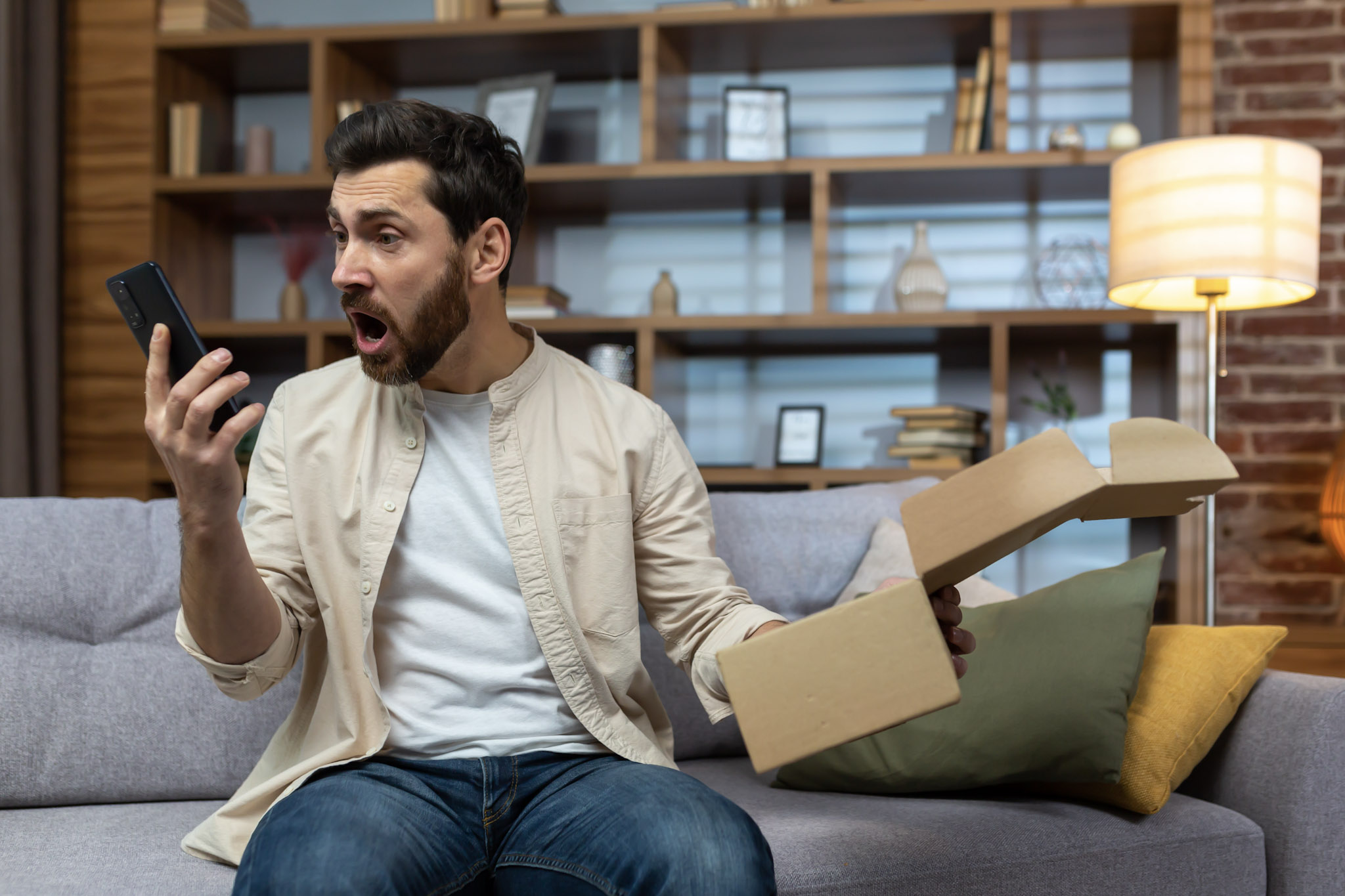 A stock image of an angry cheated man at home with a smartphone and an opened parcel box.