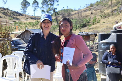 woman in WFP vest and cap stands next to Indigenous woman in pink cardigan holding a diploma in Guatemala