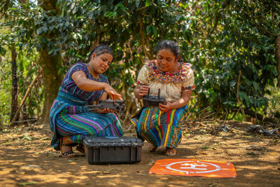 two Guatemalan Indigenous women in traditional dress crouch on the ground in a forested area operating a drone